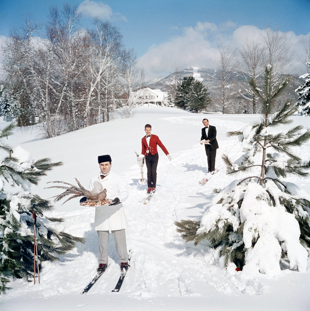 Skiing Waiters by Slim Aarons/Getty Images, 1962. Three skiing waiters on a ski slope, with the man in the foreground carrying a bird on a tray, the second man carrying a wine in an ice bucket and the third carrying a menu, Stowe, Vermont. Open Edition C-Type Photographic Print. Square orientation. Available at Electric Gallery, London, UK.