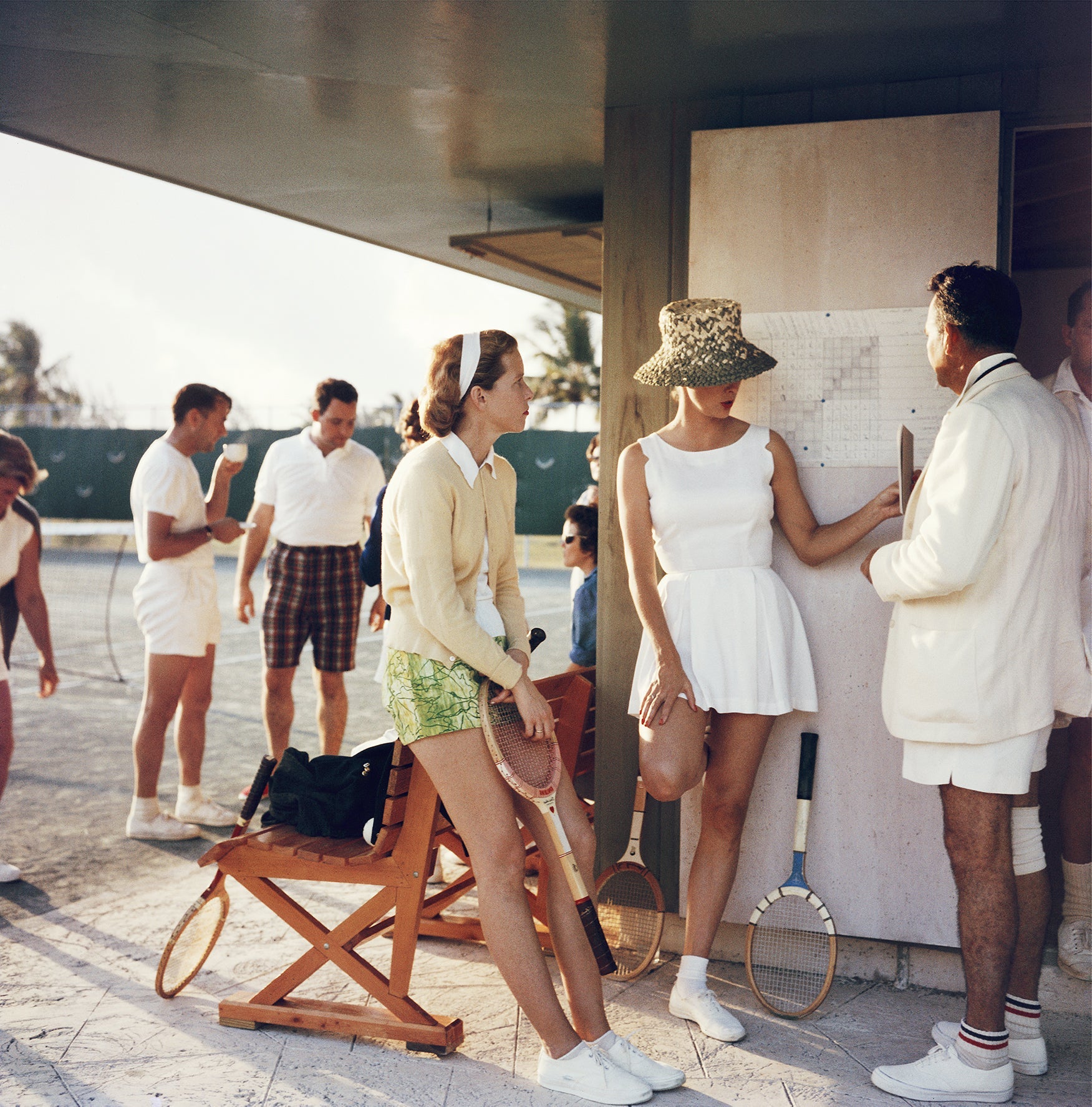 Tennis In The Bahamas by Slim Aarons/Getty Images, 1957. Two women stand talking to a man on the edge of a tennis court in the Bahamas, circa. Open Edition C-Type Photographic Print. Square orientation. Available at Electric Gallery, London, UK.