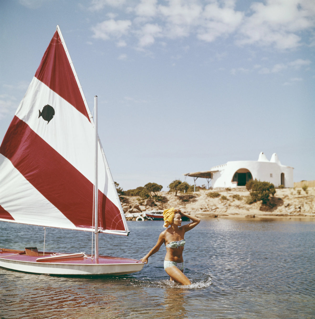 French fashion model Bettina Graziani wading in the shallow waters of Costa Smeralda, pulling a small yacht behind her, in Sardinia, Italy, 1964. Slim Aarons photograph.