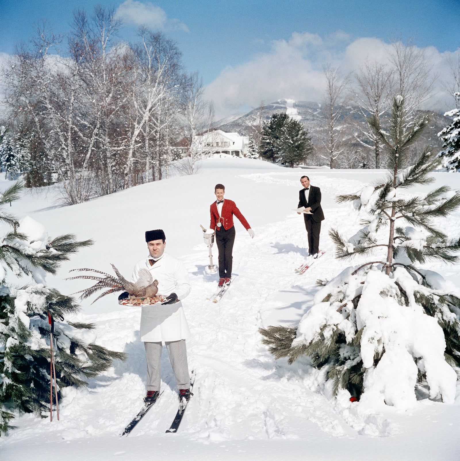 Skiing Waiters by Slim Aarons/Getty Images, 1962. Three skiing waiters on a ski slope, with the man in the foreground carrying a bird on a tray, the second man carrying a wine in an ice bucket and the third carrying a menu, Stowe, Vermont. Open Edition C-Type Photographic Print. Square orientation. Available at Electric Gallery, London, UK.