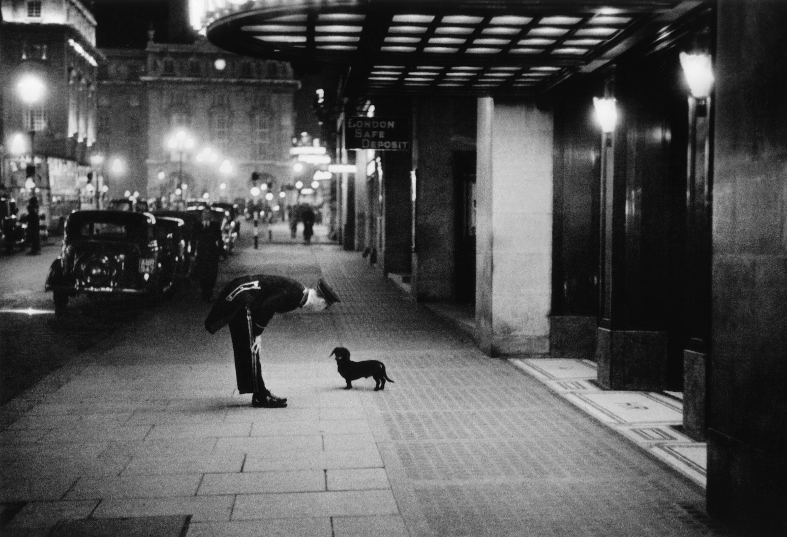 Commissionaire's Dog by Kurt Hutton/Getty Images, 1938. A hotel commissionaire talking to a small dachshund dog in Piccadilly Circus, London.. Open Edition Resin Photographic Print. Landscape orientation. Available at Electric Gallery, London, UK.